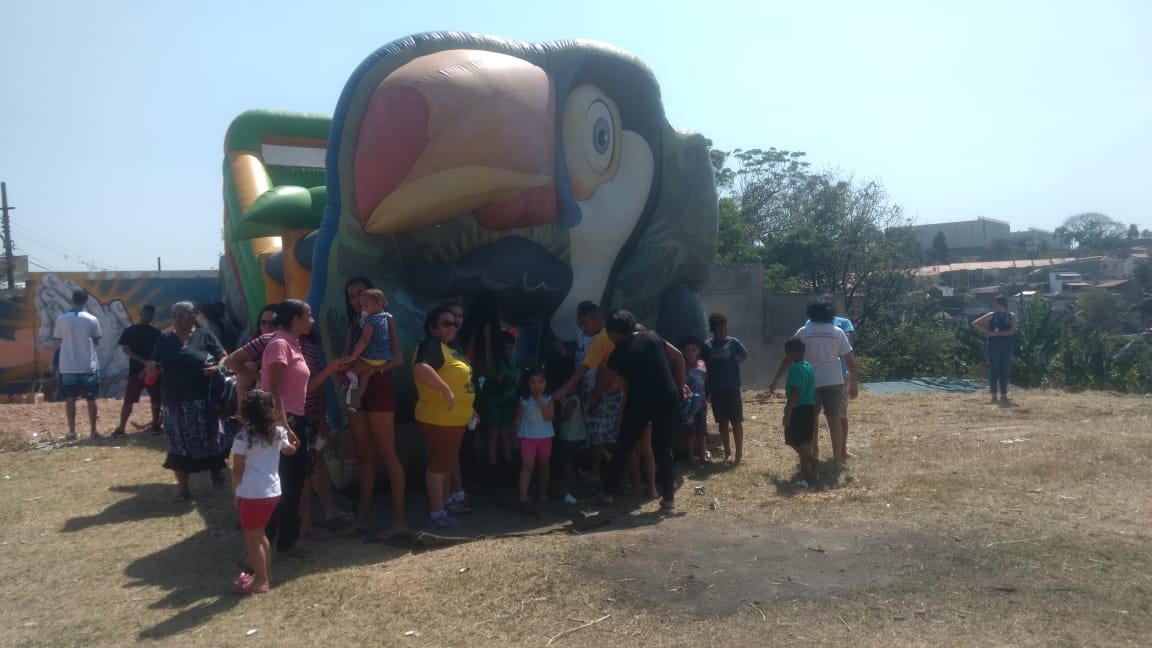 Pula-pula inflável com um desenho de um tucano em dia ensolarado e crianças em uma fila esperando para brincarem 